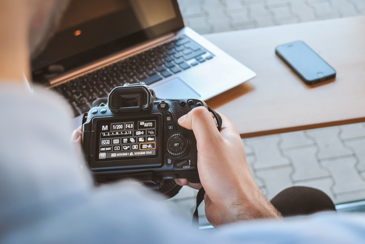 Man holding a camera with a computer and phone in the background